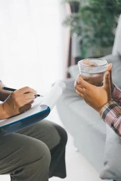 Therapist writes notes on clipboard while female client holds glass of wate.. Stock Photos