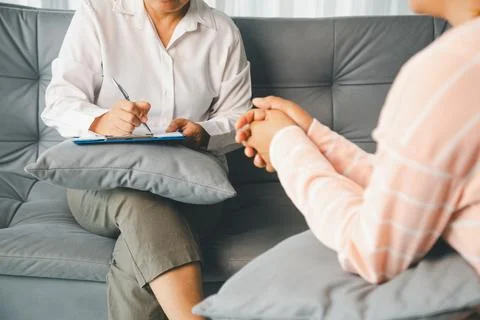 Therapist writes notes on clipboard while female client sits with clasped h.. Stock Photos