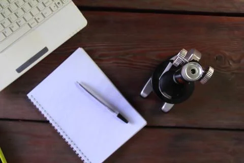 There is a microscope, a laptop and a notepad on a wooden table. Stock Photos
