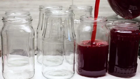 Thick dark red syrup flows smoothly from a transparent jug into a clean jar.  Stock Footage 337678758