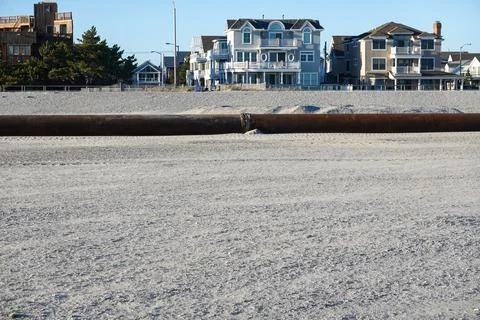 Thick rusty pipeline on a sandy beach close to a boardwalk and houses Stock Photos