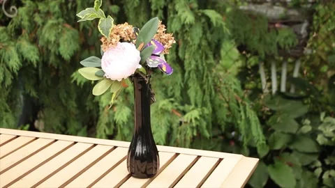 Thin dark color vase on light brown patio table with fake flowers in it Video stock 249424723