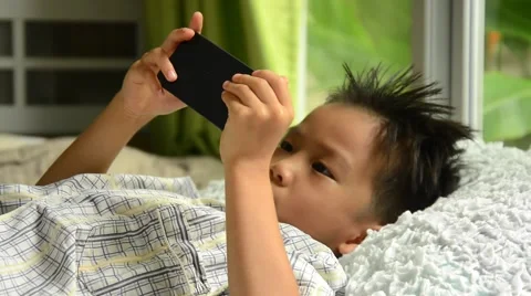 Thin focus on hand of sleepy young Asian boy using cellphone on white bed Video stock 53355875