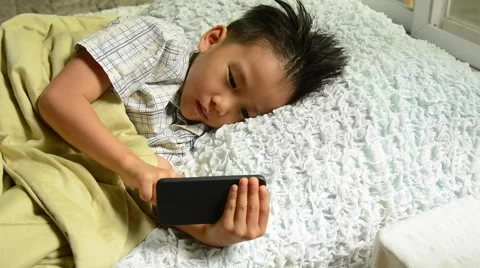 Thin focus on hand of sleepy young Asian boy using cellphone on white bed Stock Footage 53355992
