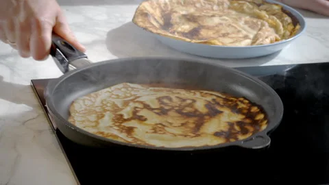 A thin pancake is being cooked in a pan, the cook moves the pan over the stove. Stock Footage 274463204