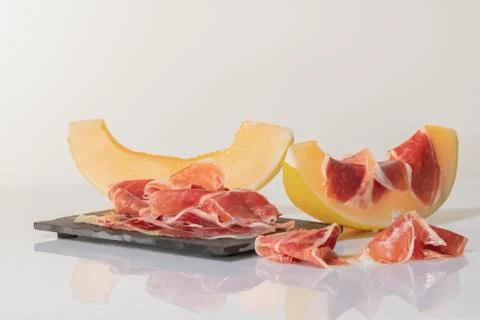 Thin slices of cured ham on a dark plate surrounded by melon slices, one of t Stock Photos