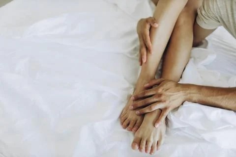 Thin smooth tender legs hug the hands of a man on white sheets. Stock Photos