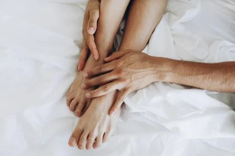 Thin smooth tender legs hug the hands of a man on white sheets. Stock Photos