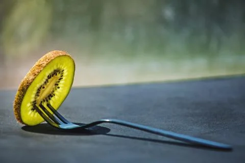 A thin transparent slice of fresh kiwi on a small black shiny dessert fork Stock Photos