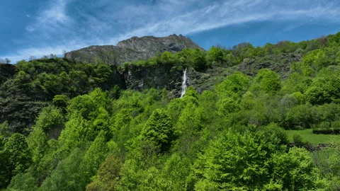 Thin waterfall cascading down rocky cliff above lush green forest and mountain Stock Footage 337951806