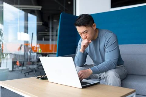 A thinking Asian man works at a computer in a modern office, a freelancer is  Foto stock