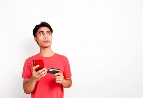 Thinking young asian man looking up while holding smartphone and credit car.. Stock Photos