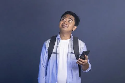 Thinking young male student with a backpack holding a smartphone to his chi.. Stock Photos