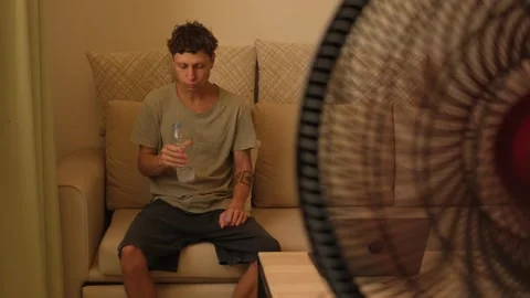 Thirsty man drinking cold water in front of electric fan during heatwave at home Stockbeeldmateriaal 346252158