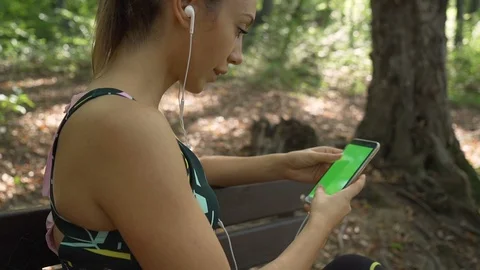 Thoughtful active girl sitting on a bench in the park, listening music from Stock Footage 117990716