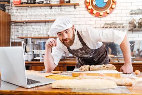 Thoughtful baker cutting bread and using laptop on kitchen Stock-Fotos