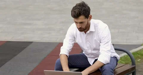 Thoughtful bearded guy sitting on bench with opened wireless laptop and doing Stock Footage 140684362