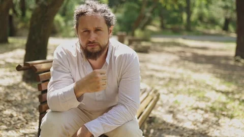 Thoughtful bearded man sitting on a park bench pointing upwards Stock Footage 323300561