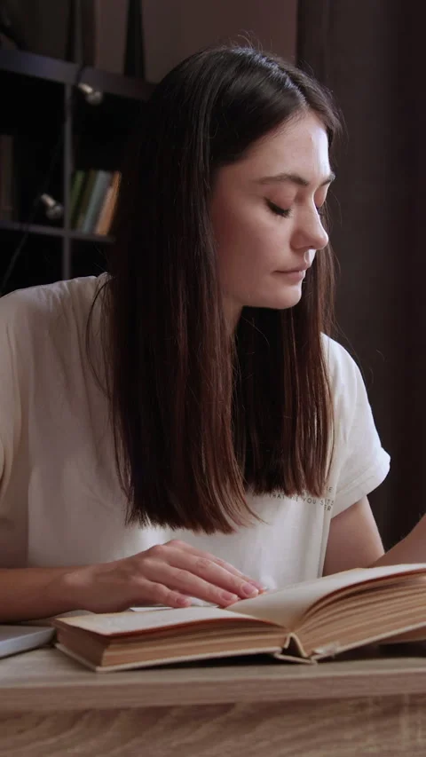 Thoughtful brunette student at an office table flips through a book in her room. Stock Footage 305409287