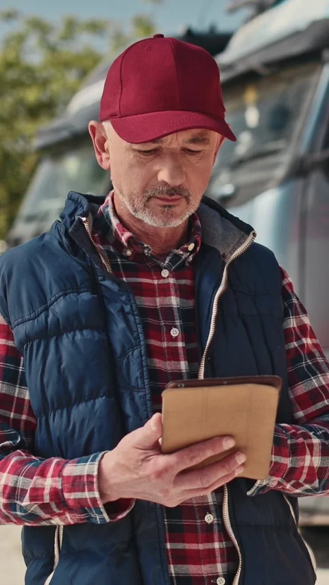 Thoughtful Caucasian man holding small tablet near parked trucks. Reviewing Stock Footage 306580607
