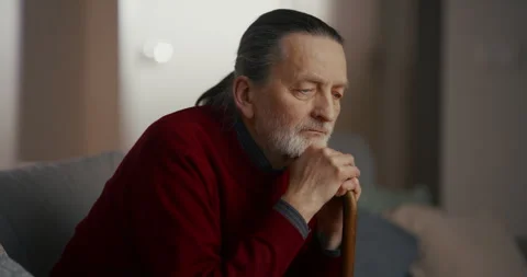 Thoughtful elderly man resting with cane near window in softly lit living room Vidéo 336278482