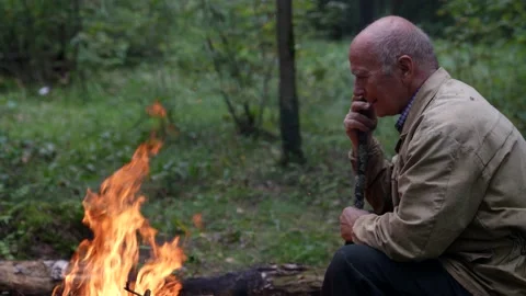 A thoughtful elderly man is sitting in the forest opposite the fire and Video stock 161005951