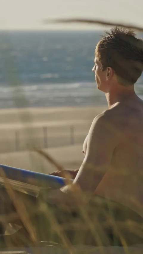 Thoughtful guy sitting by the beach at early dawn holding a surfboard Stock Footage 328670415