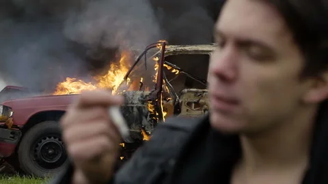 The thoughtful guy smokes against the background of his burning car, melancholy Stock Footage 95201731