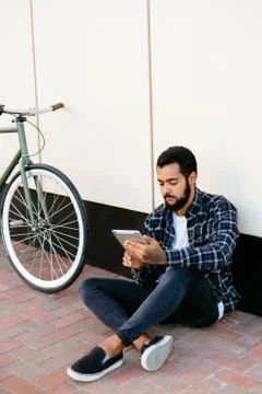 Thoughtful guy using a tablet while sitting near the bike, outdoors. Stock Photos