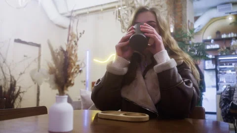 Thoughtful lady in brown shearling jacket sipping coffee in cozy caf with soft Video stock 305568481