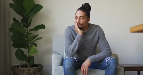Thoughtful man with dreadlocks in hair bun sitting in armchair in living room Stock Footage 337536556