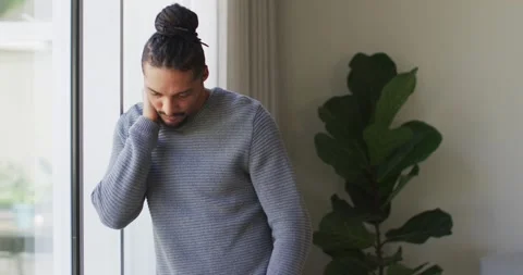 Thoughtful man with dreadlocks in hair bun looking out of window in living room Stock Footage 339650455