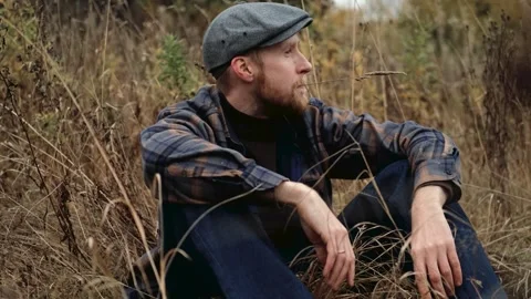 Thoughtful man in gray cap sits among tall grass, gazing into the distance Stock Footage 319962394