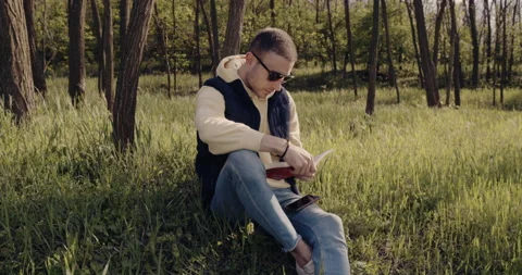 A thoughtful man sits on the grass in the woods and reads a book Video stock 133490953