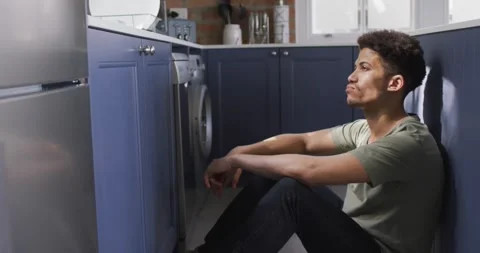 Thoughtful man sitting on floor in kitchen, leaning on countertop Stock Footage 340985759