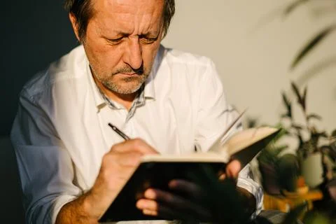 Thoughtful man sitting on a sofa, writing in notebook while concentrating d.. Stockfoto's