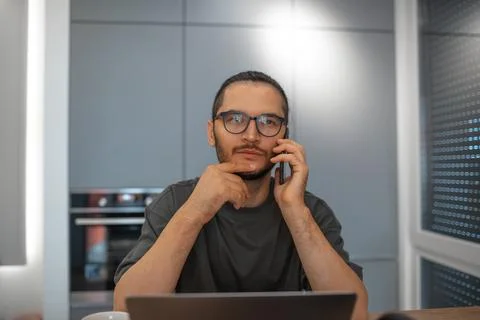 Thoughtful man talking on smartphone while working on laptop home on the ki.. Stock Photos