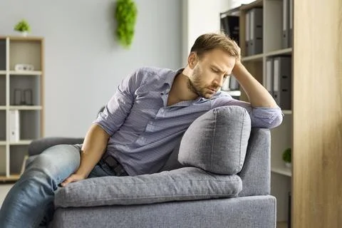 Thoughtful man by window on sofa, stressed and anxious, reflecting life problems 写真素材