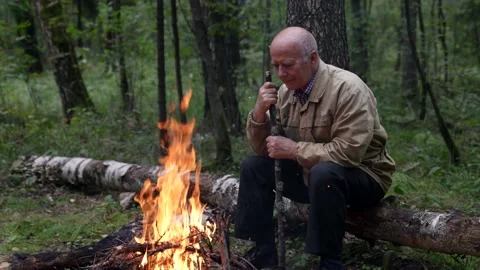 A thoughtful old man sits alone by a campfire in the woods late at night, looks 스톡 동영상 166078111
