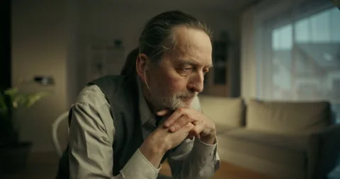Thoughtful older man sitting alone by a window in a softly lit living room Stock Footage 336286158