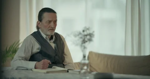 Thoughtful older man sitting at a table in a bright living room Stock Footage 336287455