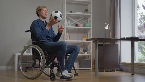 Thoughtful smiling disabled young man in wheelchair with soccer ball indoors at Vídeo Stock 164932480