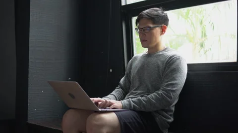 Thoughtful young asian man sit on window with laptop computer  thinking and t Stock Footage 158794351