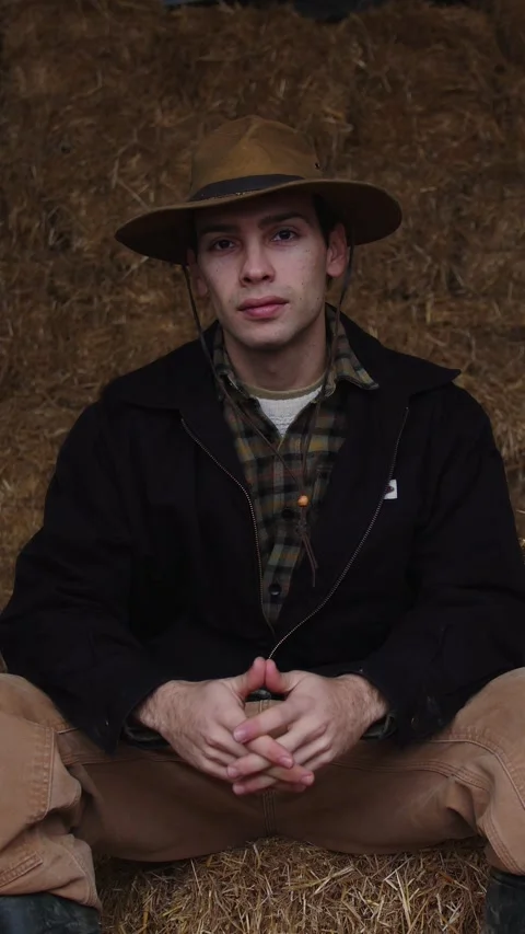 Thoughtful Young Farmer Seated in Barn, Contemplating, Surrounded by Hay with a Stock Footage 265944705