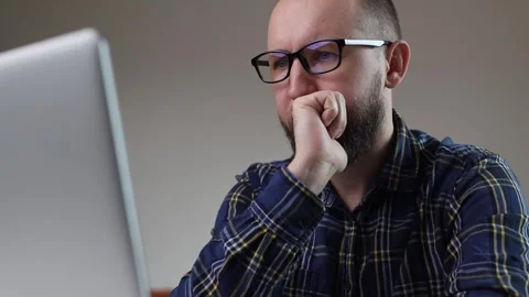 Thoughtful young man with a beard in glasses sits at a laptop Stock-Footage 243372034