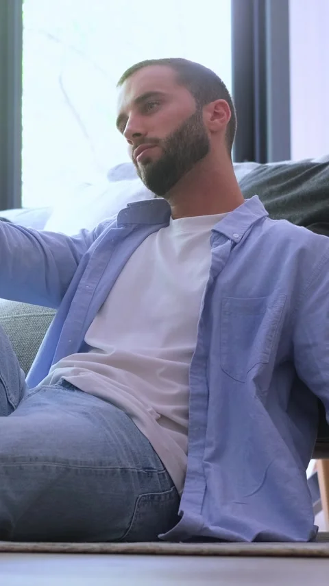 Thoughtful young man with a beard sitting on the floor, leaning against the Vídeos de archivo 344757142