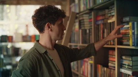 Thoughtful young man chooses book from library shelf, enjoying reading hobby Stock Footage 342731227