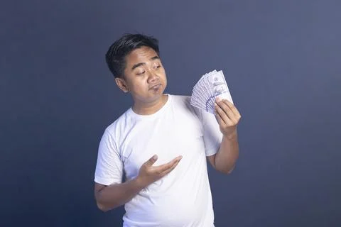 Thoughtful young man gesturing toward a fan of dollar bills in his hand, co.. Stock Photos