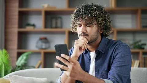 Thoughtful young man holding mobile phone sitting on sofa in living room looking Vídeos de archivo 334225039