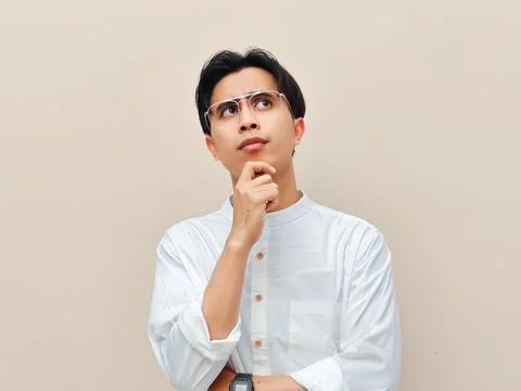 Thoughtful young man looking up and thinking with hand on chin against a be.. Stock Photos